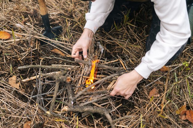 Primo piano ritagliato di un turista irriconoscibile che mette l'accensione sul fuoco ardente per riscaldarsi e cucinare il cibo all'aperto in una giornata fredda e nuvolosa. Concetto di bushcraft, campeggio e sopravvivenza in natura.