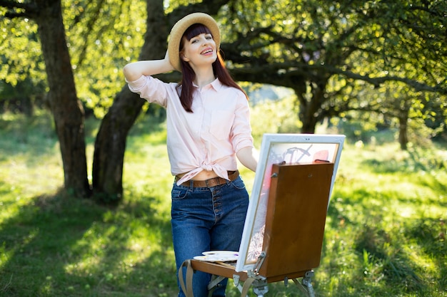 Primo piano ritratto all'aperto di giovane affascinante donna caucasica con cappello di paglia, camicia e pantaloni di jeans, in posa in un giardino, mentre si diverte a dipingere un quadro su tela.