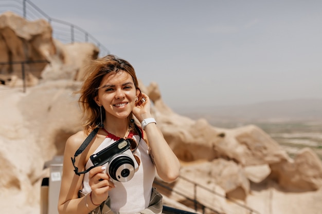 Primo piano ritratto all'aperto di una ragazza sorridente felice che tiene in mano una fotocamera retrò e cammina tra vecchie rocce alla luce del sole