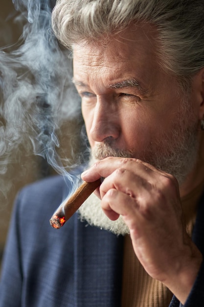 Primo piano ritratto di brutale uomo maturo dai capelli grigi che guarda lontano mentre fuma sigaro