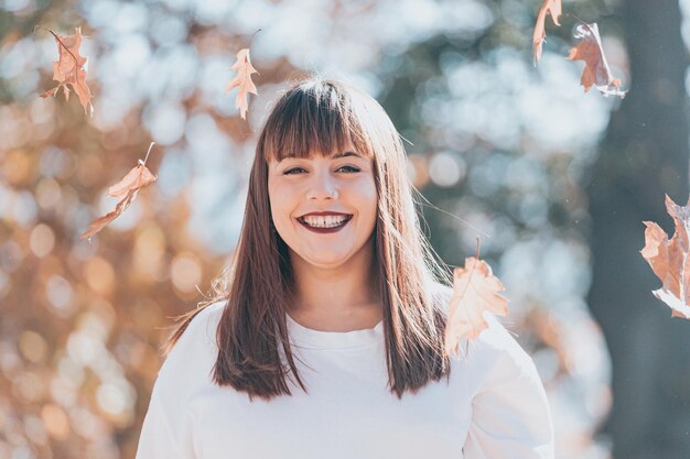 Primo piano ritratto di giovane donna felice che si diverte nella foresta autunnale. Ritratto di donna che lancia foglie d'autunno. La ragazza felice in camicia bianca e jeans blu sta giocando con la foglia, sta esaminando la macchina fotografica e sta sorridendo.