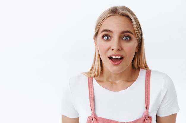 Primo piano scioccato e impressionato ragazza bionda con gli occhi azzurri in tuta rosa, t-shirt, bocca aperta e sguardo affascinato scoperto notizie incredibili, in piedi sfondo bianco ansimando per l'interesse