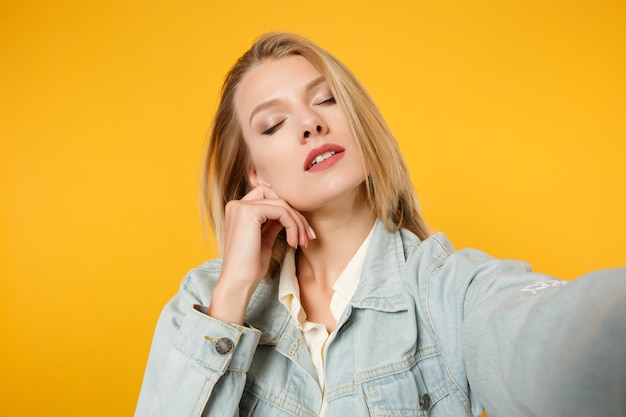 Primo piano selfie di tenera giovane donna in abiti casual denim mettere la mano sul mento, tenendo gli occhi chiusi isolati su sfondo giallo arancione. Concetto di stile di vita delle persone. Mock up spazio di copia.