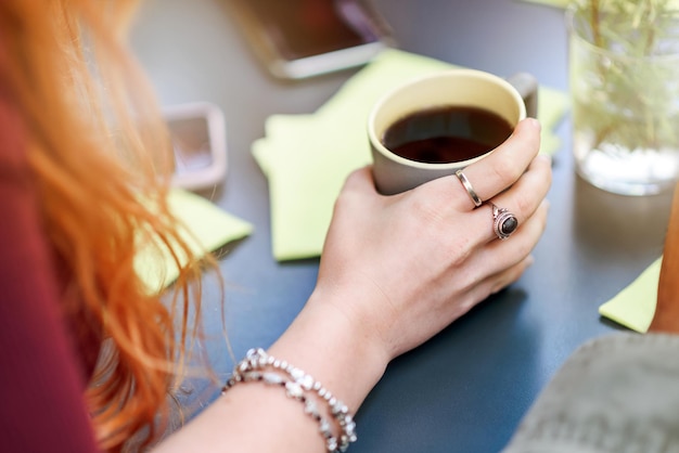 Primo piano su una mano femminile che tiene una tazza americana di caffè nero appena fatto in una tazza di ceramica in una vista da sopra la spalla seduti a un tavolo