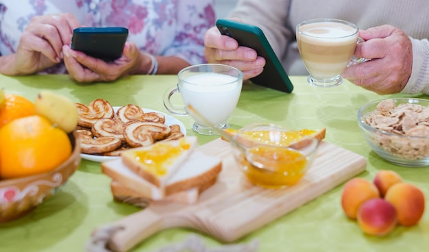 Primo piano sulle mani degli anziani adulti che tengono utilizzando gli smartphone mentre si fa colazione a tavola a casa