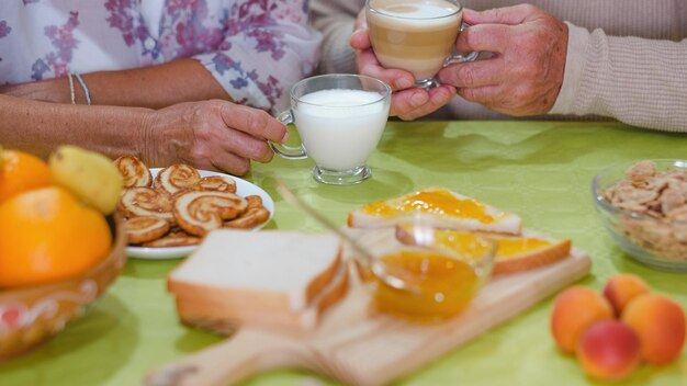 Primo piano sulle mani dell'anziano adulto che tengono una tazza di latte e cappuccino mentre si fa colazione a tavola