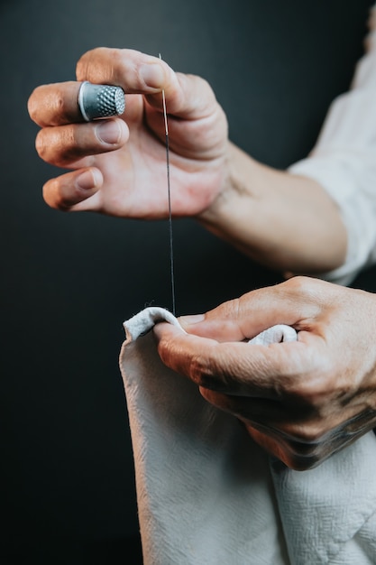 Primo piano sulle mani della donna per cucire ago e filo. Vecchia donna che lavora con le mani sprecate. Sarto che cuce un po' di tessuto. Dettagli, poca luce, lunatico