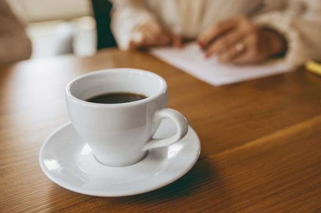 Primo piano tazza di caffè davanti a mani femminili che scrivono note durante riunioni creative, discussioni, progetti di lavoro in ufficio. Concetto di finanza, affari, potere della ragazza, inclusione, diversità, femminismo.