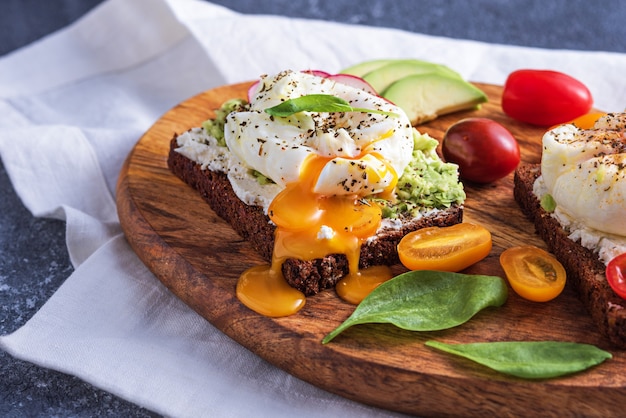 Primo piano toast con uovo in camicia, ricotta, avocado e verdure su tavola di legno su carta da cucina bianca, sana colazione rurale concept