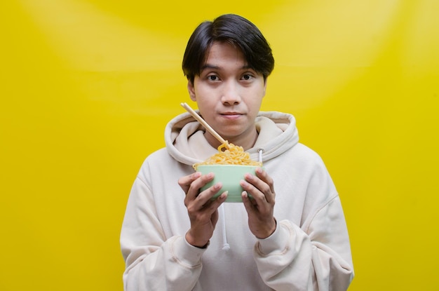 Primo piano un uomo asiatico in una felpa con cappuccio beige mangia spaghetti istantanei usando le bacchette e una ciotola