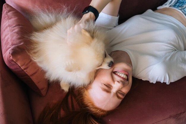 Primo piano vista dall'alto del simpatico cane bianco Spitz adorabile che lecca il viso di una giovane donna ridente sdraiata su un comodo divano. Attraente donna rossa che trascorre il tempo libero con un soffice cagnolino a casa.