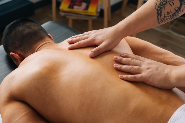 Primo piano vista dall'alto di un massaggiatore maschio irriconoscibile con forti mani tatuate che massaggiano la schiena di uno sportivo muscolare irriconoscibile sdraiato sullo stomaco