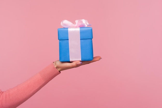 Primo piano vista laterale mano femminile che tiene e mostra confezione regalo blu con fiocco d'argento regali e bonus Studio indoor girato isolato su sfondo rosa