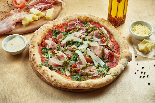 Primo piano vista su appetitosa pizza con prosciutto, pesto, parmigiano, rucola e pomodori secchi sul tavolo di legno in un ristorante. Cucina italiana.