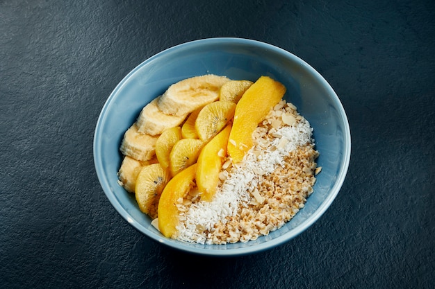 Primo piano vista sulla sana colazione: farina d'avena con cocco, mango, banane e kiwi giallo in una ciotola blu su un tavolo nero.