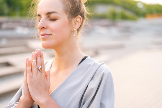 Primo piano volto di calma attraente giovane donna che pratica yoga eseguendo namaste posa con gli occhi chiusi fuori nel parco cittadino Bella donna serena meditando e praticando yoga all'aperto da solo