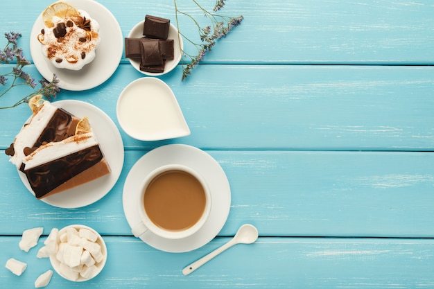 Priorità bassa del caffè del mattino. Tazza da cappuccino di confine, barattolo di latte, cioccolato, piccole torte e zucchero raffinato su tavolo in legno rustico blu, spazio copia, vista dall'alto