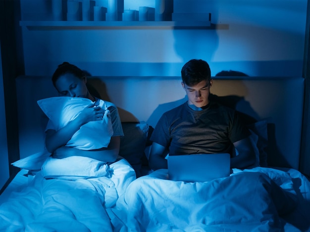 Problema di insonnia Relazione di coppia Dipendenza da tecnologia digitale Dipendenza da gadget Uomo occupato che digita laptop di notte vicino a sonnecchiare sul cuscino interno camera da letto oscura donna