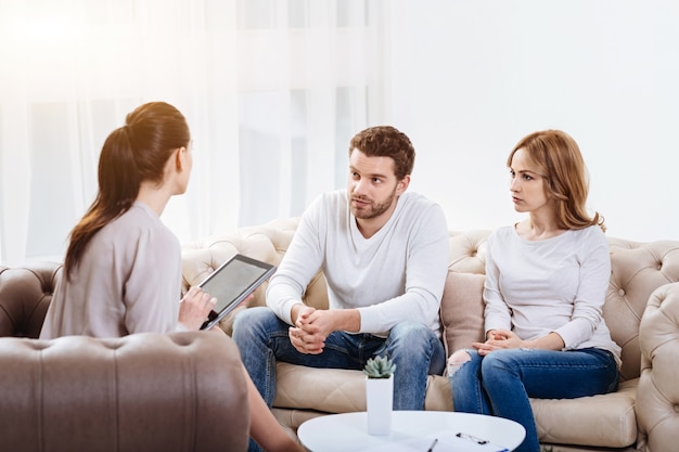 Problemi nella relazione. Cupa bella giovane coppia seduta sul divano e guardando lo psicologo mentre discute dei loro problemi