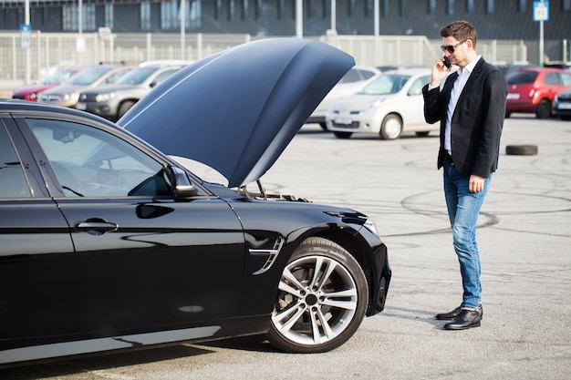 Problemi sulla strada. Uomo con l'automobile rotta che rivolge allo smartphone