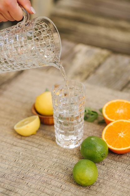 Procedimenti di versare acqua fredda in un bicchiere alto di agrumi freschi sul tavolo, arancia, limone e lime