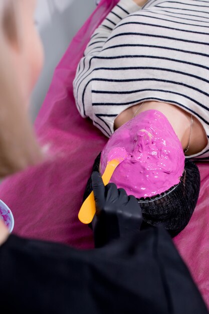 Processo di applicazione della maschera di alginato. Mani femminili di una spatola della tenuta dell'estetista. La procedura per la cura della pelle nel salone di bellezza. Donna sdraiata su un divano nell'ufficio di un'estetista.