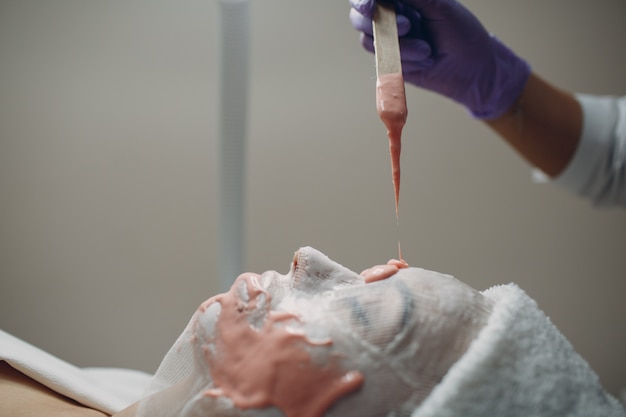 Processo di applicazione della maschera facciale in alginato. Mani femminili di una spatola della tenuta dell'estetista. Donna sdraiata su un divano in ufficio di cosmetologo. Cura della pelle del viso nel salone di bellezza spa.