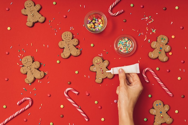 Processo di preparazione del biscotto di panpepato di Natale con omino di pan di zenzero, glassa, granelli di zucchero e bastoncini di zucchero su uno sfondo rosso della tavola. Carta da parati di Capodanno piatta.