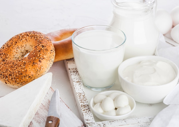 Prodotti lattiero-caseari freschi in scatola di legno d'annata sulla tavola bianca. Vaso e bicchiere di latte, ciotola di panna acida e formaggio. Bagel appena sfornati.