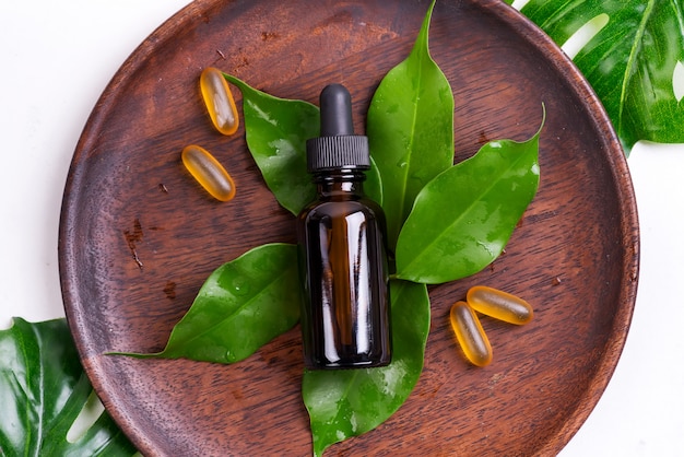 Prodotti naturali di bellezza con le capsule di gel di Omega 3 e siero in bottiglie di vetro, foglie verdi sul piatto di legno su bianco