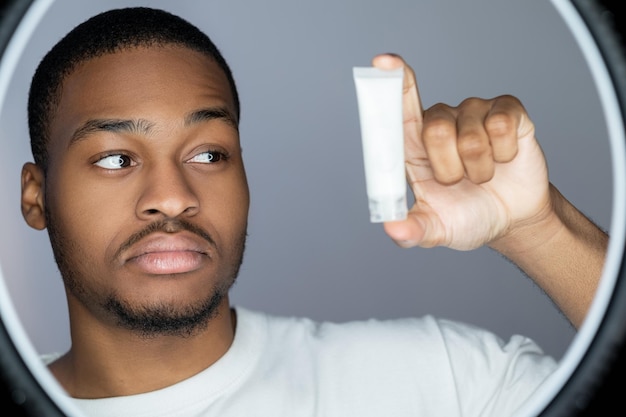 Prodotto cosmetico per il trattamento del viso per la cura della pelle dell'uomo confuso ragazzo africano dubbioso che guarda il mockup bianco