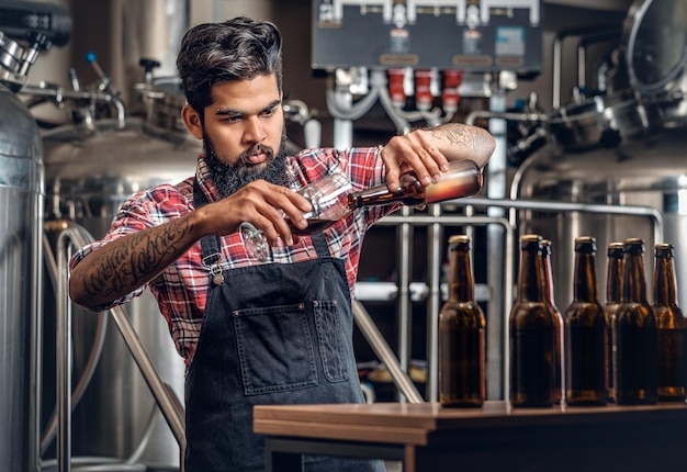 Produttore maschio indiano barbuto e tatuato hipster che assaggia e presenta birra artigianale nel microbirrificio.