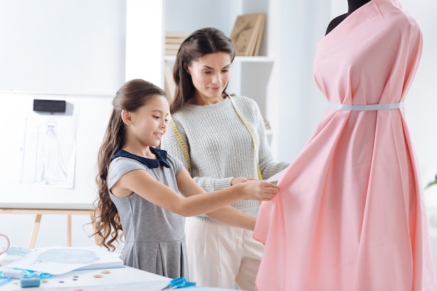 Professione futura. Ragazza piacevole piacevole creativa che sta vicino a sua madre e che sorride mentre progetta un vestito