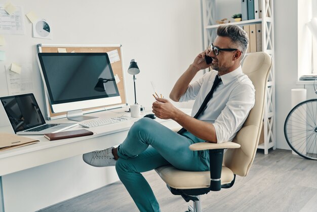 Professionista di successo. Bel giovane in camicia e cravatta che parla allo smartphone e sorride mentre è seduto in ufficio