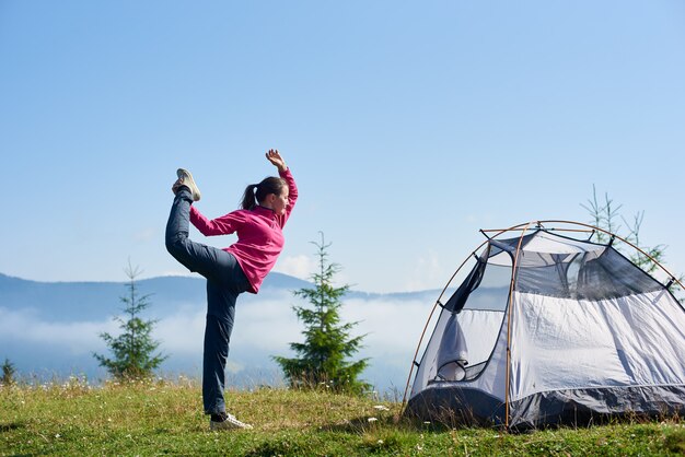 Profili il punto di vista di giovane ragazza turistica esile che sta su una gamba nella posa dell'yoga sulla valle erbosa verde alla tenda turistica sotto bello cielo blu sulle montagne nebbiose