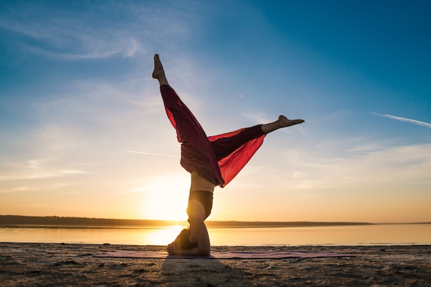 Profili la donna sulla spiaggia al tramonto facendo asana di yoga. Allenamento mattutino di riscaldamento elasticizzato naturale