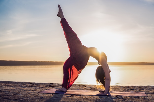 Profili la donna sulla spiaggia al tramonto facendo asana di yoga. Allenamento mattutino di riscaldamento elasticizzato naturale