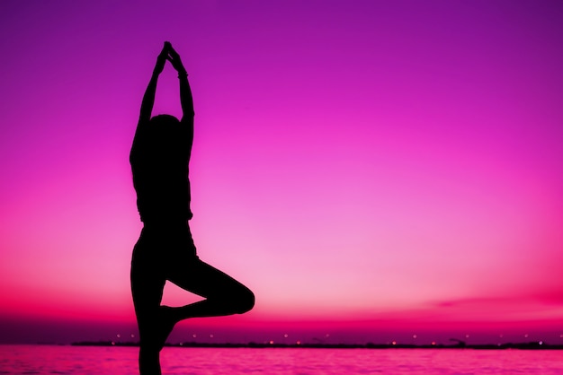 Profili la pratica meditating della donna di posa di yoga sui precedenti del tramonto della spiaggia