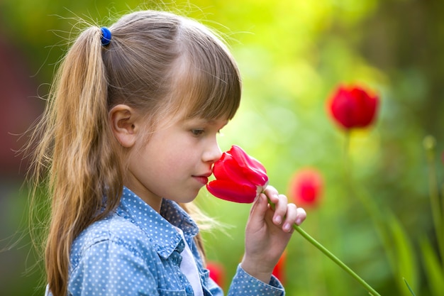 Profilo della ragazza sorridente graziosa sveglia del bambino con gli occhi grigi e capelli lunghi che odorano il fiore rosso luminoso del tulipano sul giardino vago del bokeh di verde di estate soleggiata vaga.