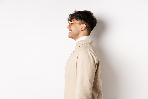 Profilo di bell'uomo caucasico in occhiali, guardando lo spazio vuoto lasciato con un sorriso allegro, in piedi sul muro bianco.