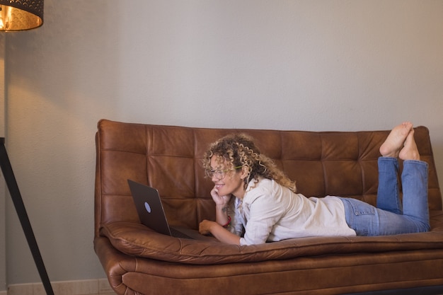 Profilo di donna sorridente in occhiali e capelli ricci che guarda contenuti multimediali utilizzando il computer portatile su un comodo divano in pelle a casa. Donna felice che guarda un film sul portatile a casa