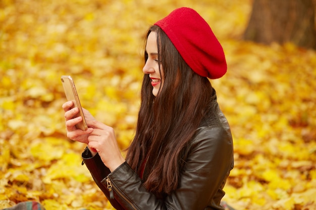 Profilo di giovane attraente ragazza carina abiti berretto rosso e giacca di pelle, tenendo il telefono intelligente davanti a lei