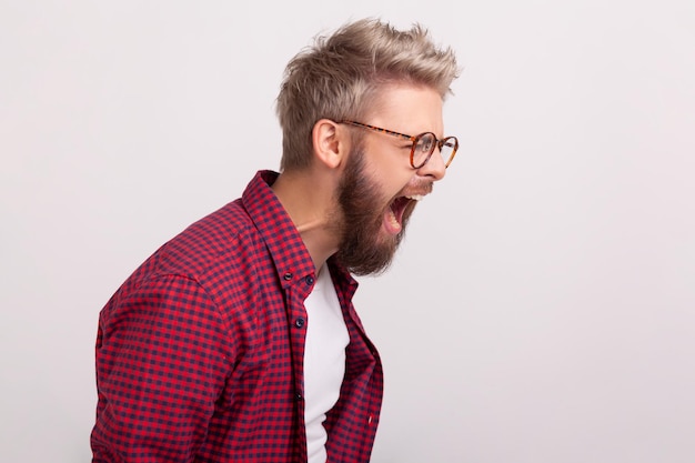 Profilo di uomo barbuto furioso in occhiali e camicia rossa a scacchi che urla esprimendo rabbia e irritazione Colpo in studio indoor isolato su sfondo grigio