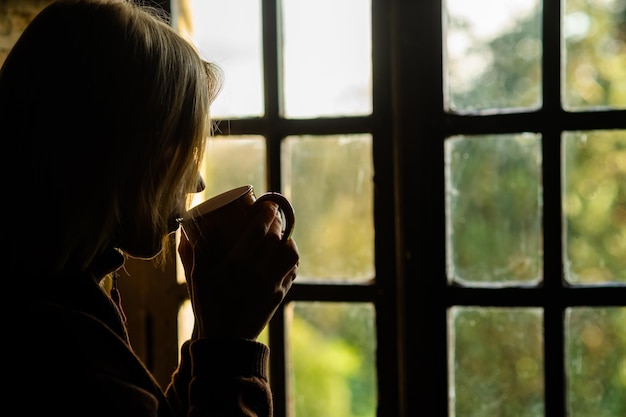 Profilo scuro di una donna con una tazza in mano vicino alla finestra Caffè del mattino