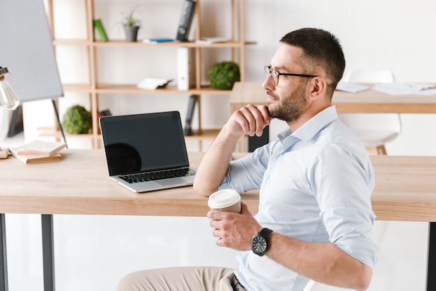 Profilo uomo ufficio diligente 30s in camicia bianca seduto a tavola e bere caffè da asporto, mentre si lavora al computer portatile nel centro business