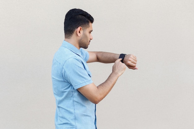 Profilo vista laterale ritratto di attento bel giovane uomo barbuto in camicia blu in piedi e usando il suo orologio intelligente, toccando lo schermo. colpo dello studio dell'interno isolato sul fondo beige chiaro della parete.