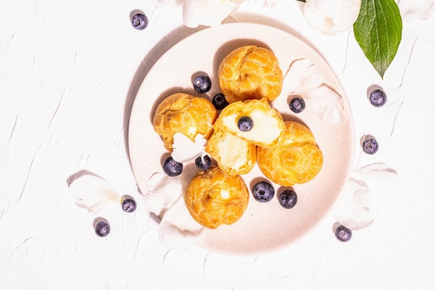 Profiteroles ai mirtilli. Torte alla crema, Eclair, bignè. Luce dura moderna, ombra scura. Peonie bianche su fondo in gesso, vista dall'alto