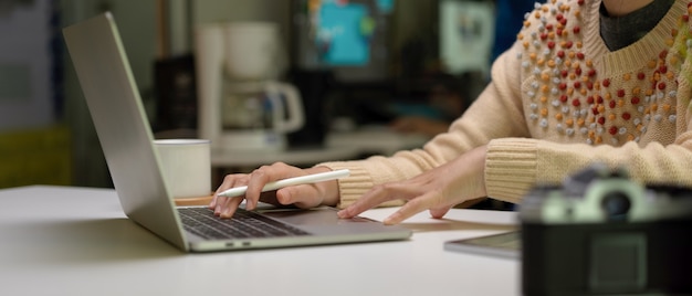 Progettista femminile che per mezzo del computer portatile sulla scrivania bianca con la tazza della macchina fotografica, della compressa e di caffè