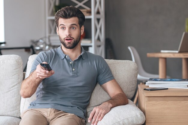 Programma televisivo scioccante. Bel giovane che tiene il telecomando e sembra sorpreso mentre guarda la TV sul divano di casa