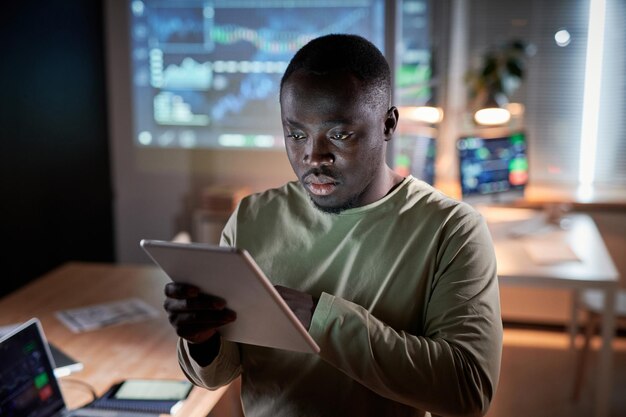 Programmatore africano che esamina il nuovo software su tablet digitale che lavora in ufficio fino a tarda sera